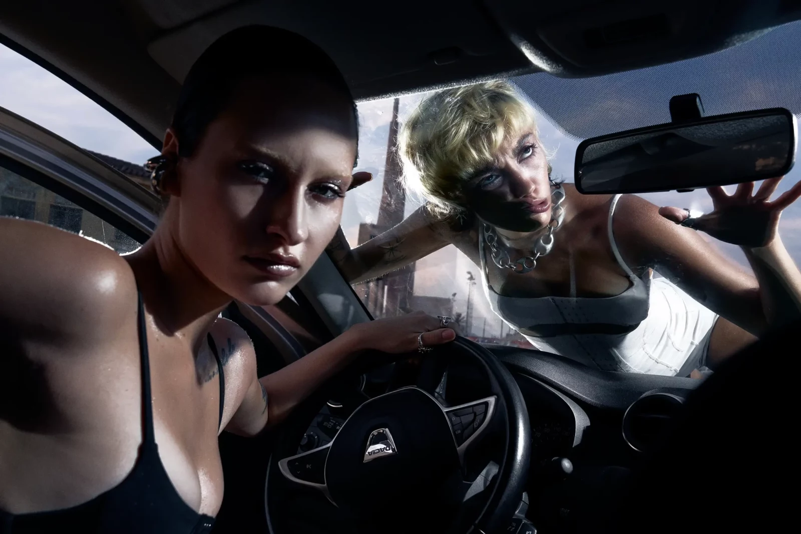 Fashion model in black leans over a car steering wheel while a short-haired blonde model looks through the window.