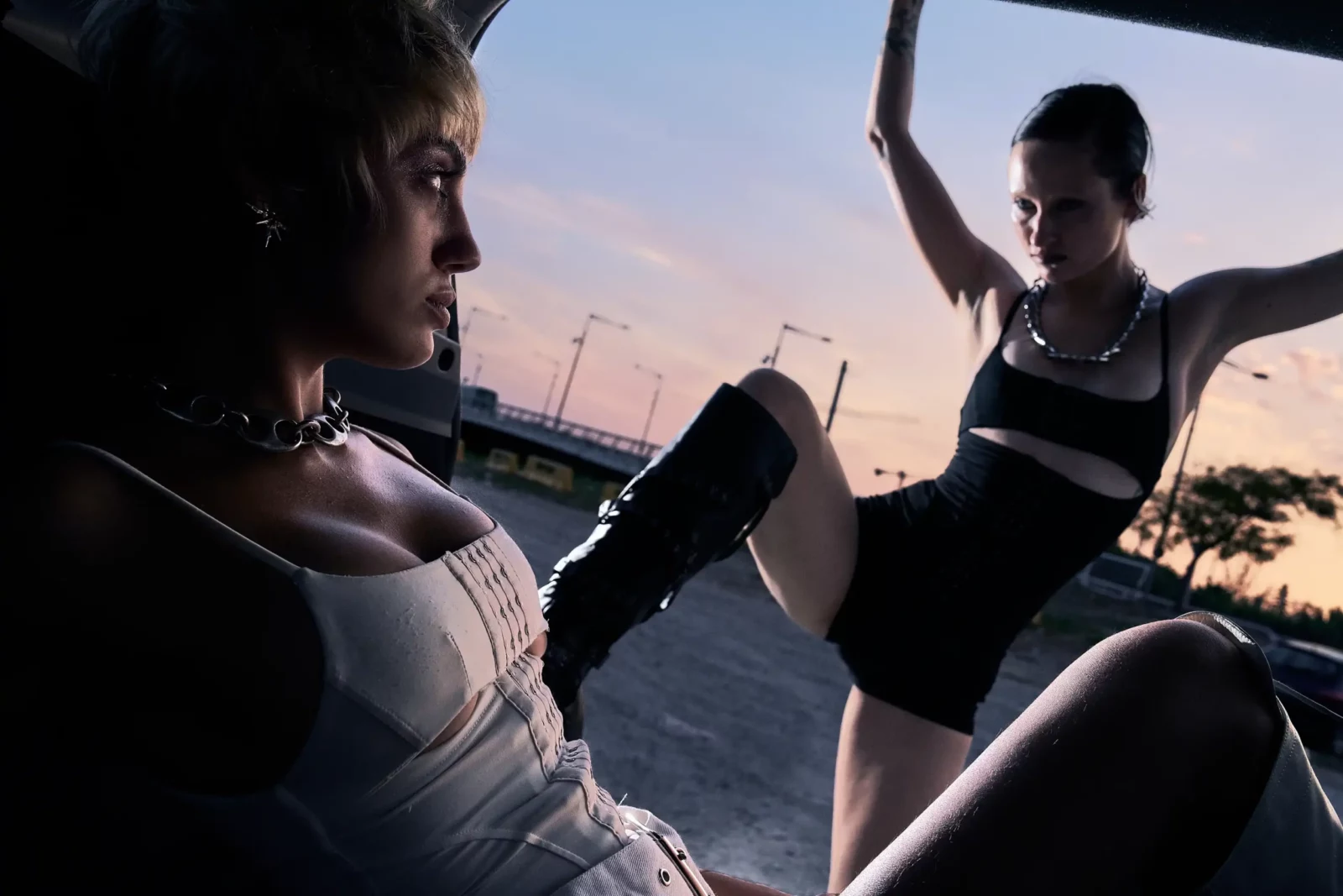 Two fashion models interact inside an open car at sunset, one wearing a white top and the other black.