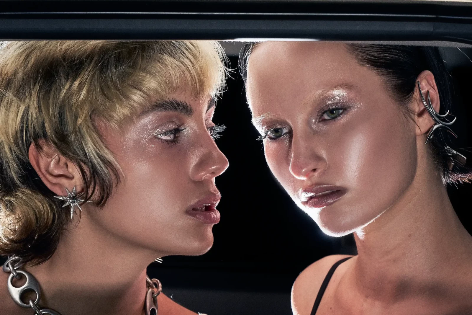 Close portrait of two models framed by a car window, wearing bold eye makeup and statement jewelry.
