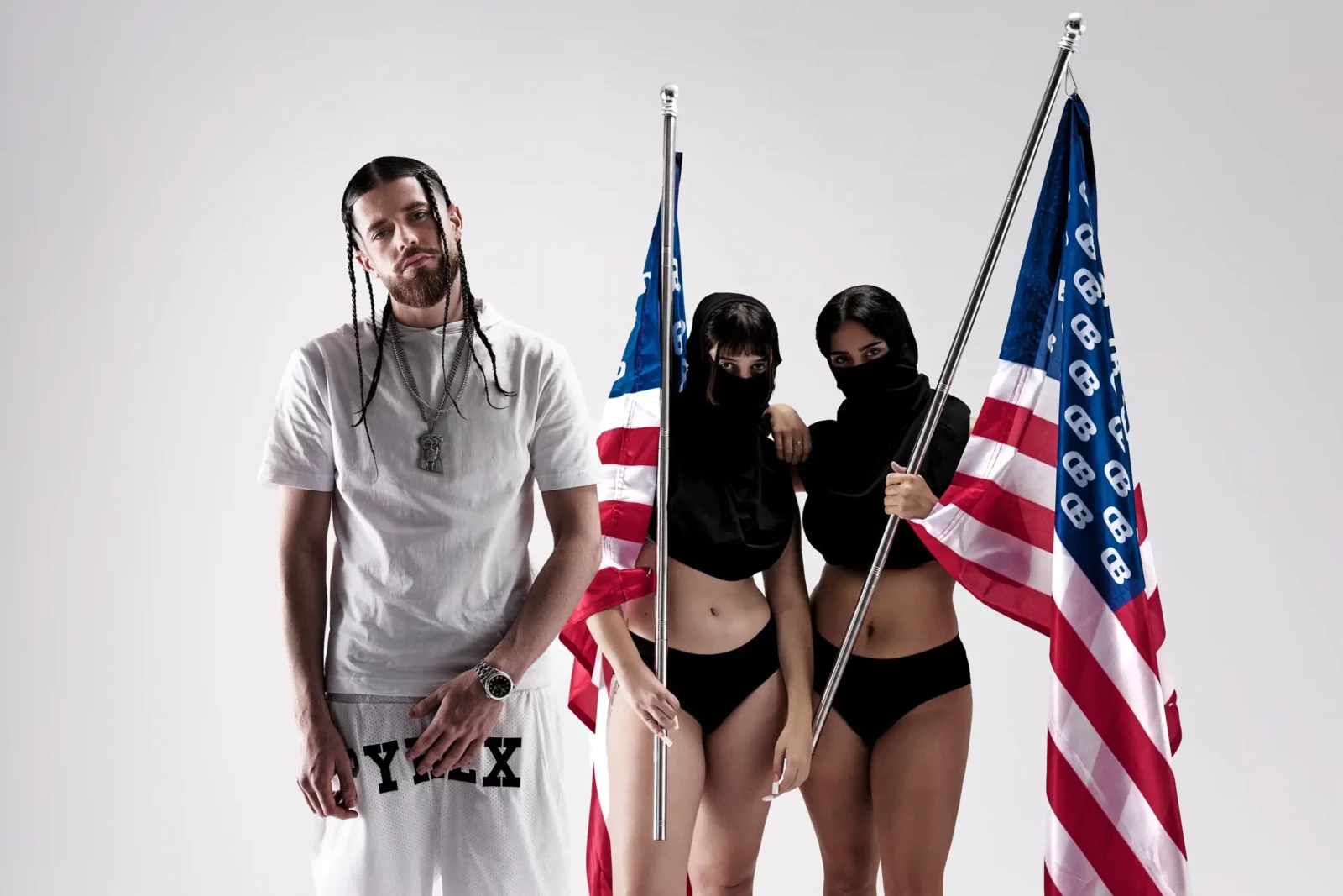Long-haired musician in a white shirt standing between two masked women holding American flags.