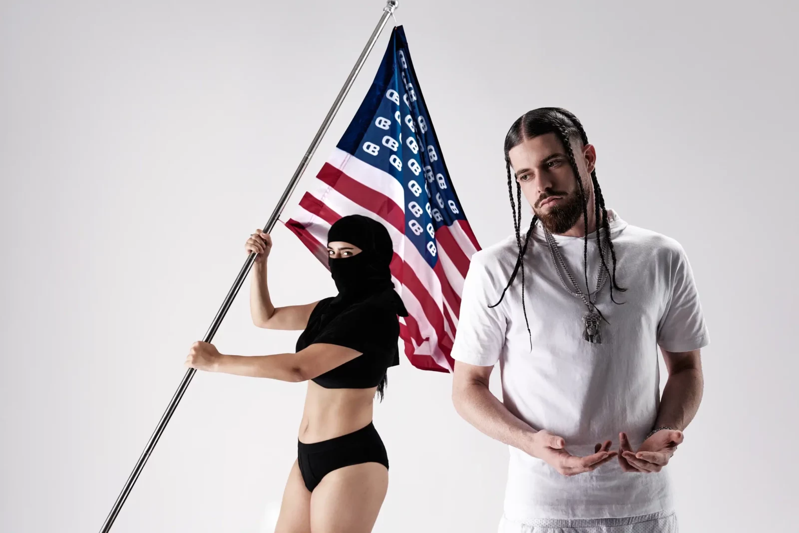 Musician in a white shirt posing with a masked woman holding an American flag against a white backdrop.