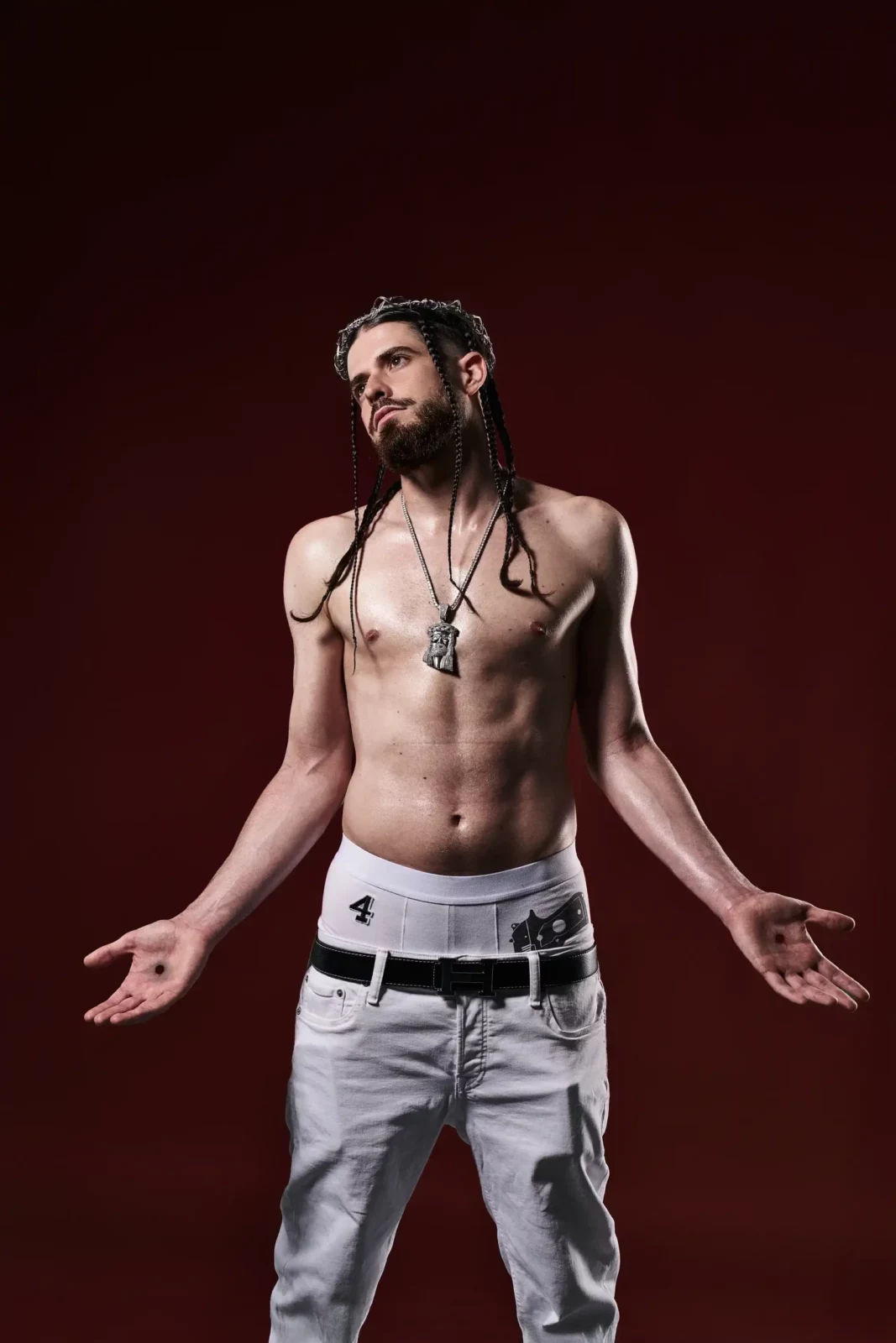 Shirtless musician with long braided hair posing against a deep red studio background.