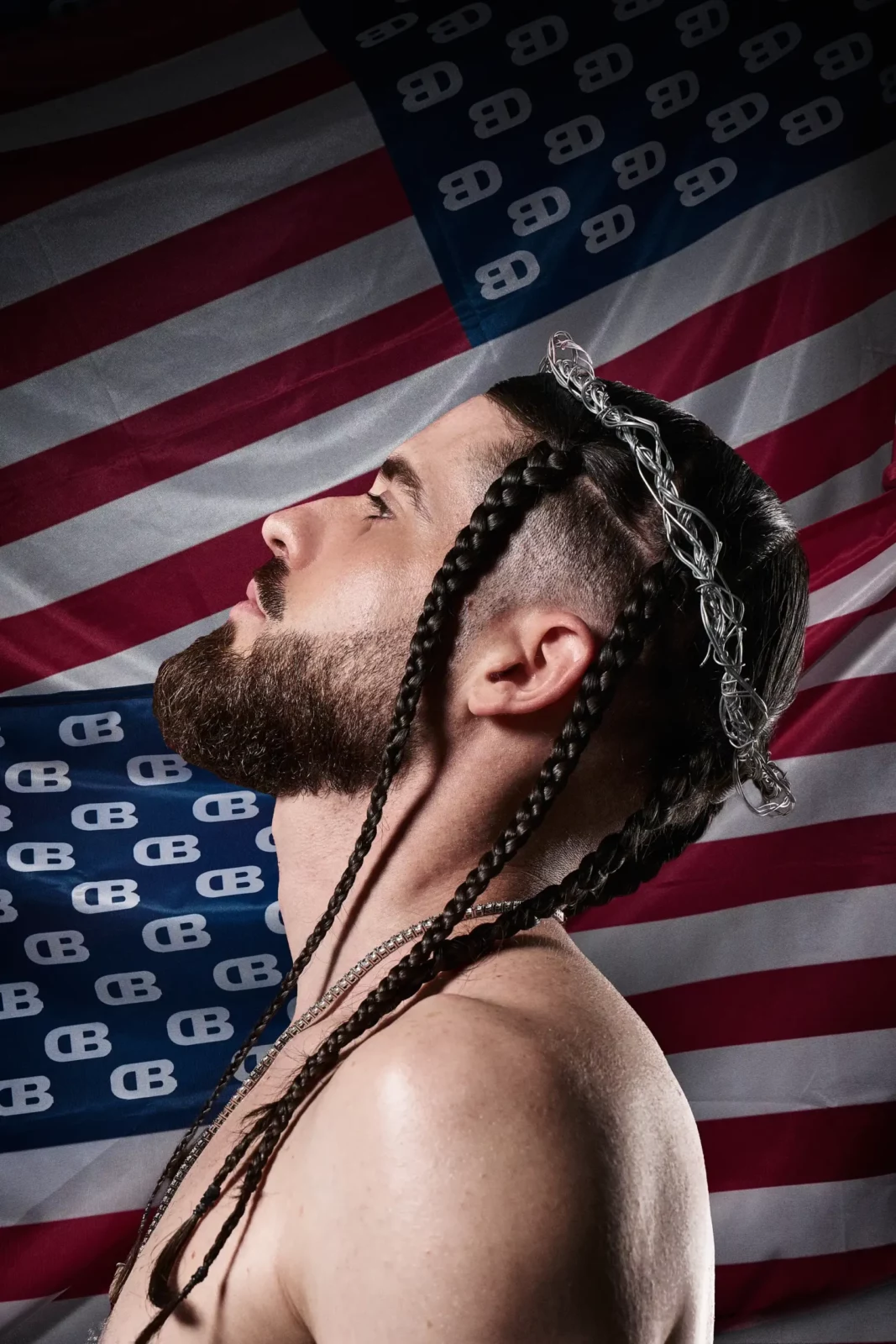 Side-profile close-up of a long-haired musician with braids against a large American flag.