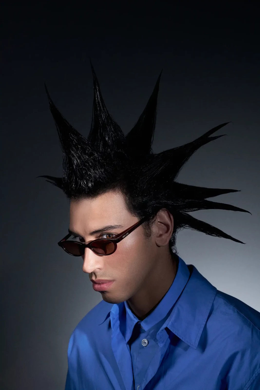 Close-up studio portrait of a model with spiked black hair and narrow black sunglasses.