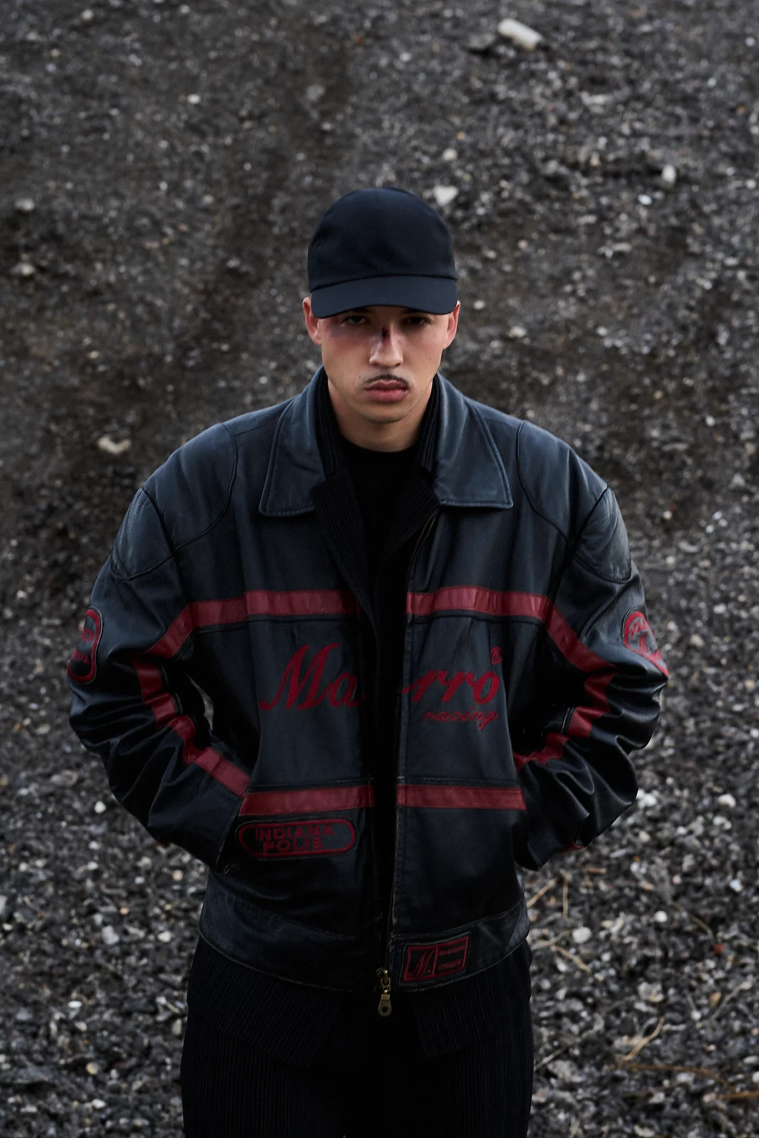 Front-facing portrait of Pole in a black baseball cap and dark striped jacket standing against gravel.