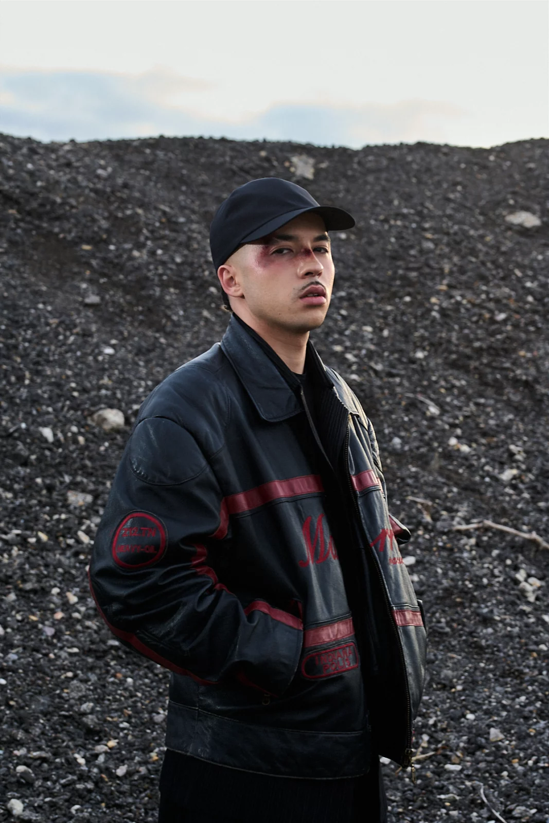 Pole standing in profile in a black cap and oversized dark jacket with red stripes against a gravel landscape.