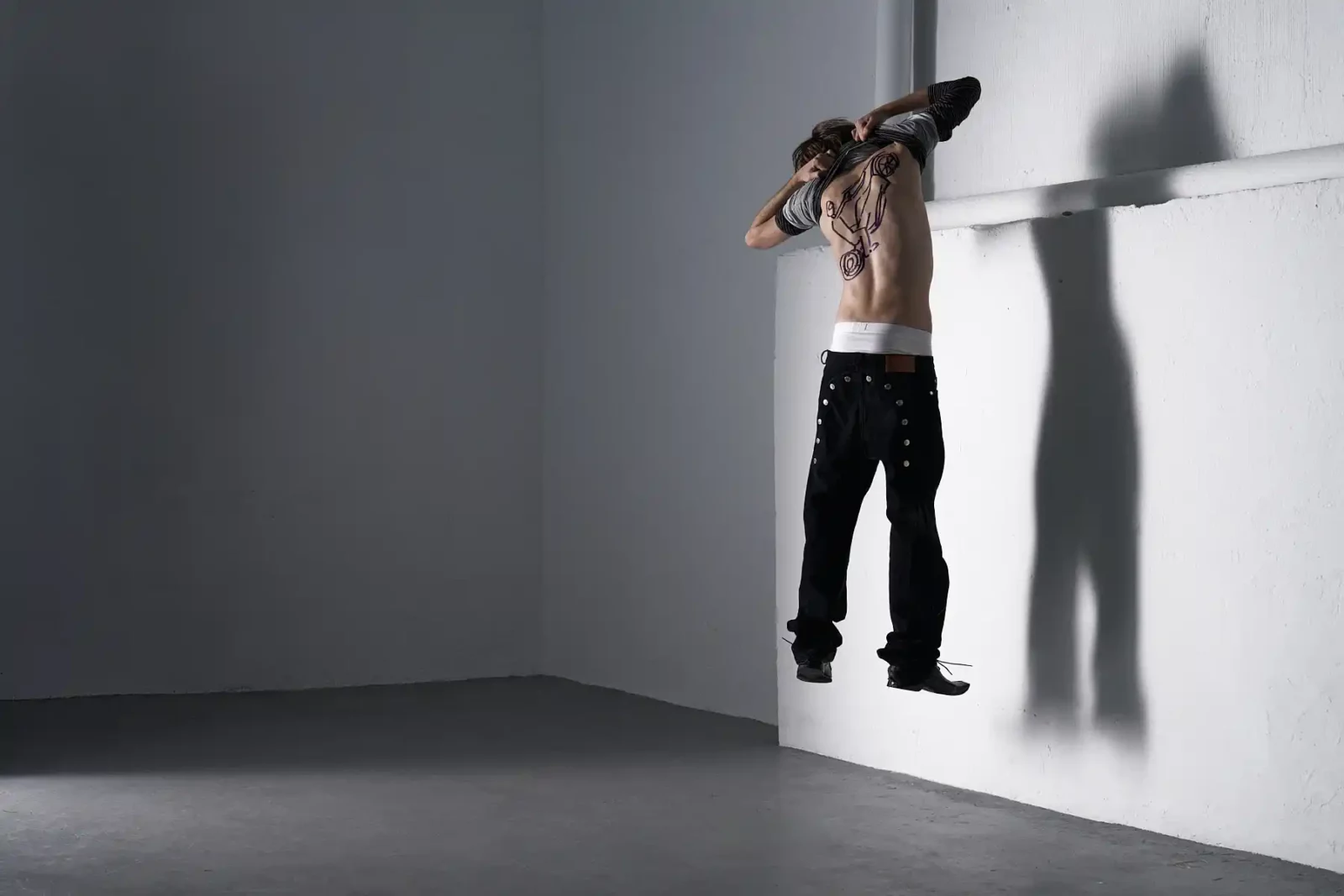 Minimalist studio photograph of a shirtless model appearing to float beside a white wall, casting a strong shadow.