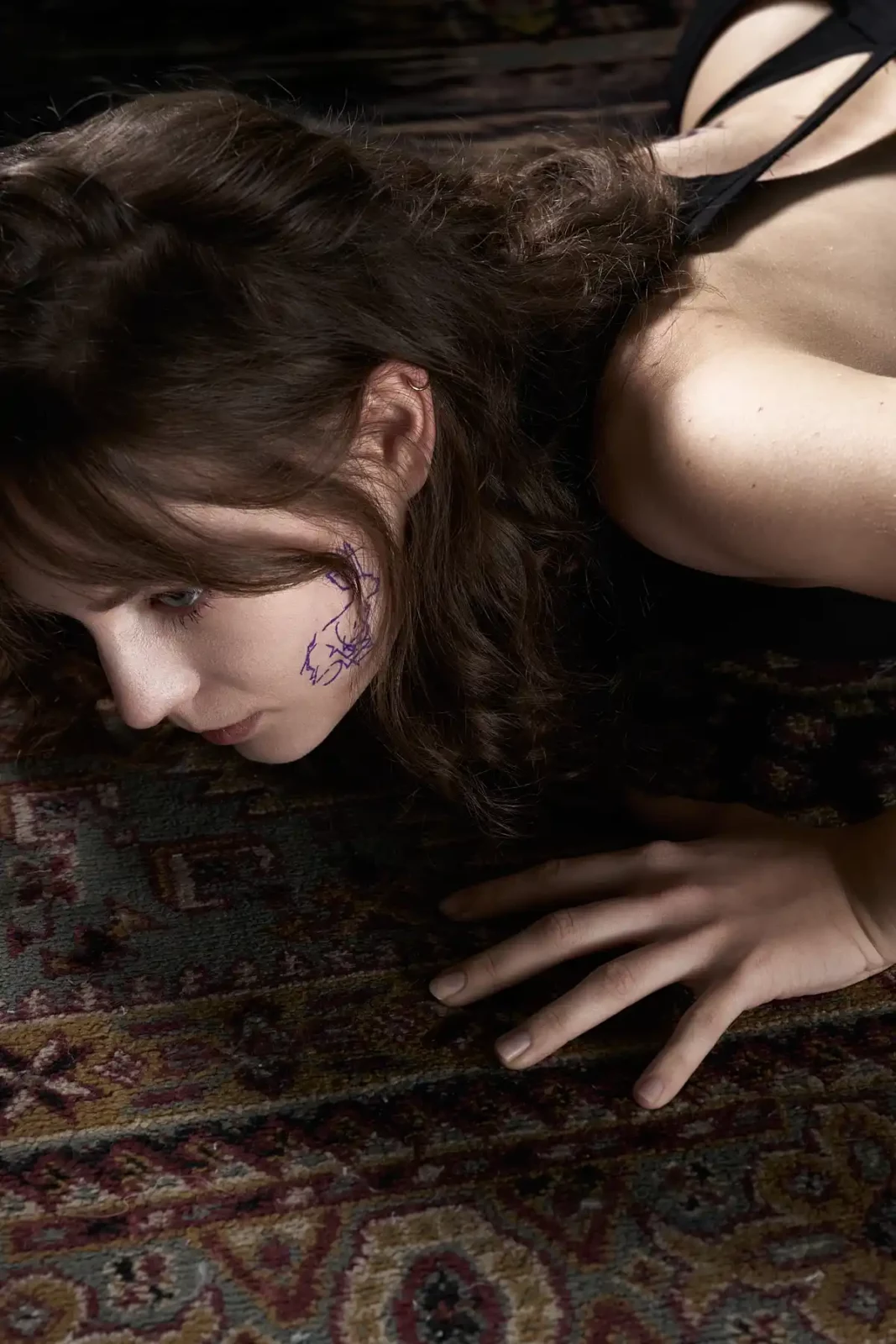 Close-up portrait of a dark-haired model lying on a patterned rug with a purple face tattoo design.