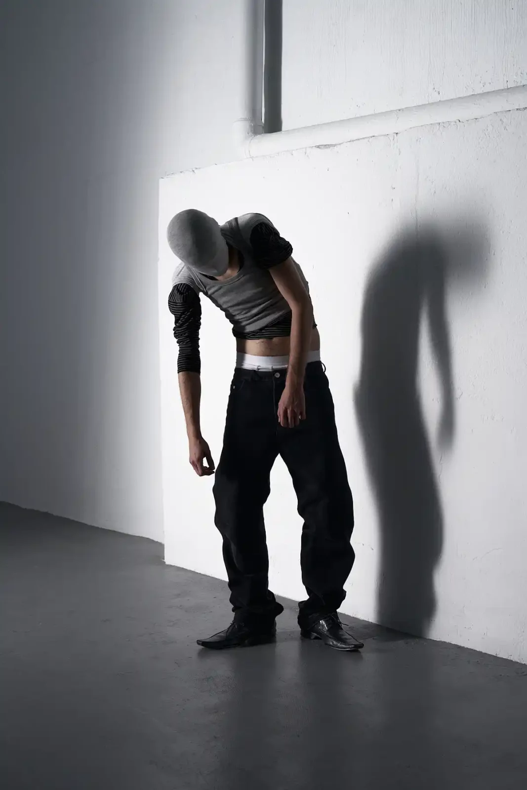 Shirtless model with dark trousers bending forward beside a white studio wall and strong cast shadow.
