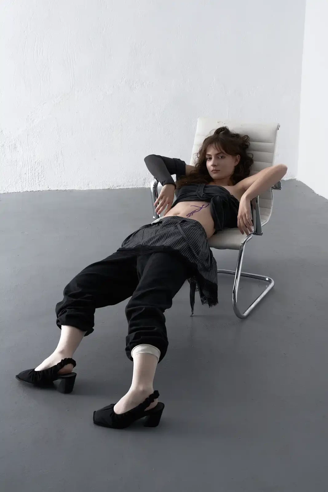 Model reclining with arms behind the head in a chair wearing black clothing and low heels.