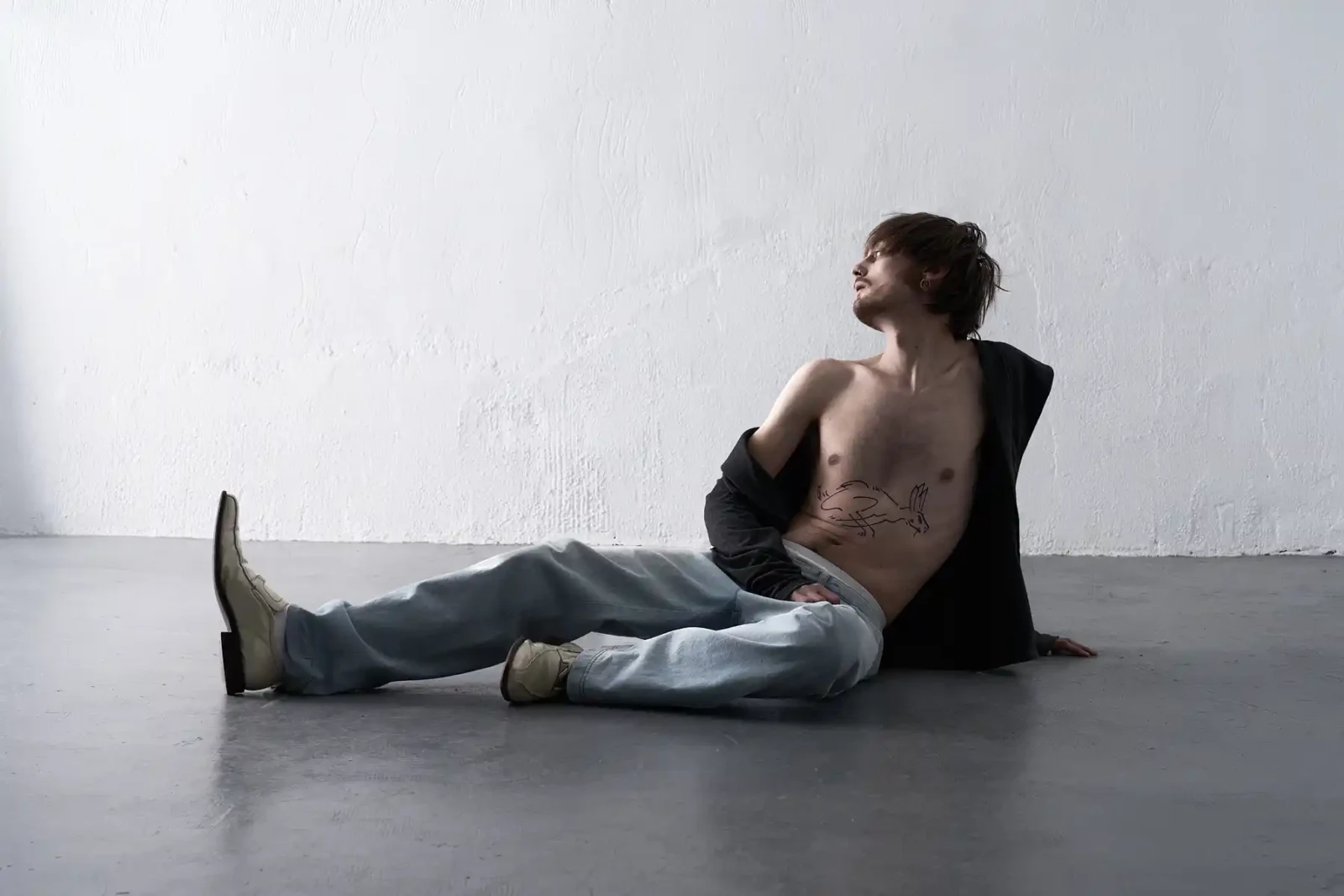 Shirtless male model in loose light jeans sitting on a gray floor against a white studio wall.
