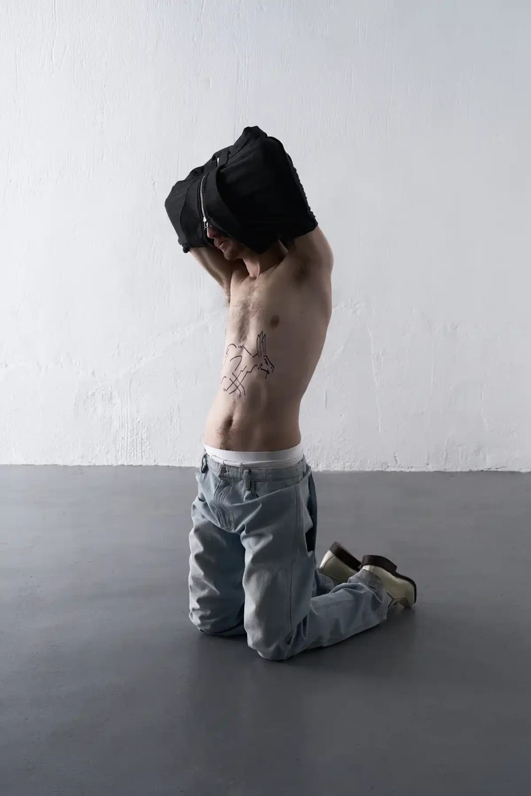 Shirtless model wearing pale blue jeans and a black cloth hood kneeling in a white studio.