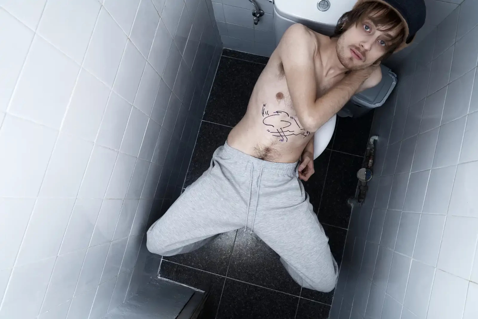 Overhead portrait of a shirtless tattooed male model wearing gray sweatpants in a narrow tiled restroom.