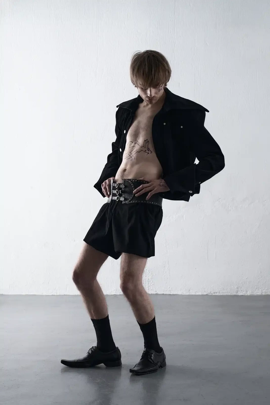 Male model in a cropped black jacket and black shorts posing against a light studio wall.