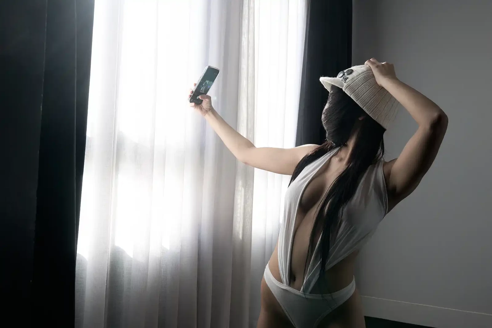 Woman wearing a white cap and pale bodysuit taking a selfie next to a bright hotel window.