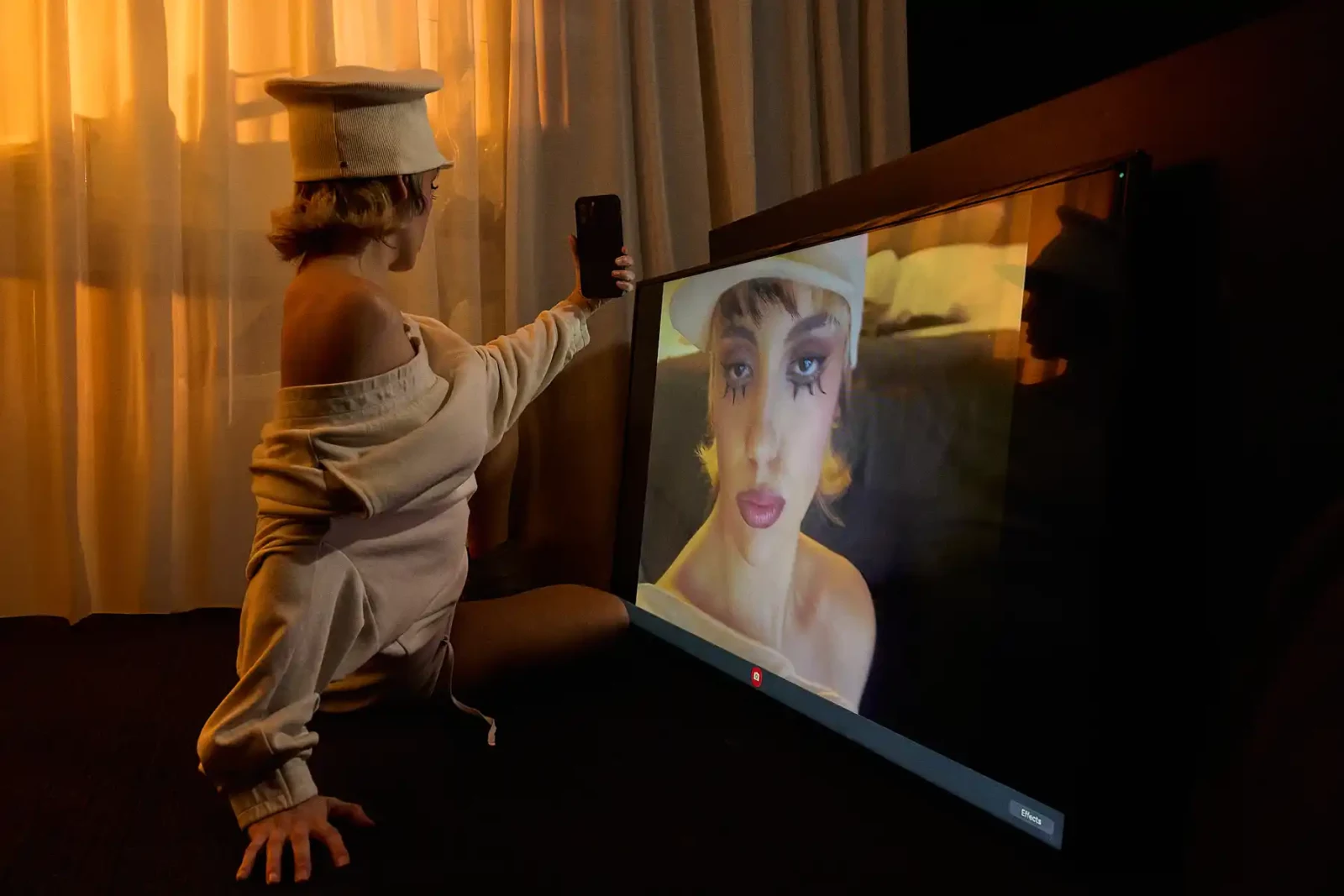 Model in a white cap and off-the-shoulder top sitting beside a screen showing a beauty portrait.