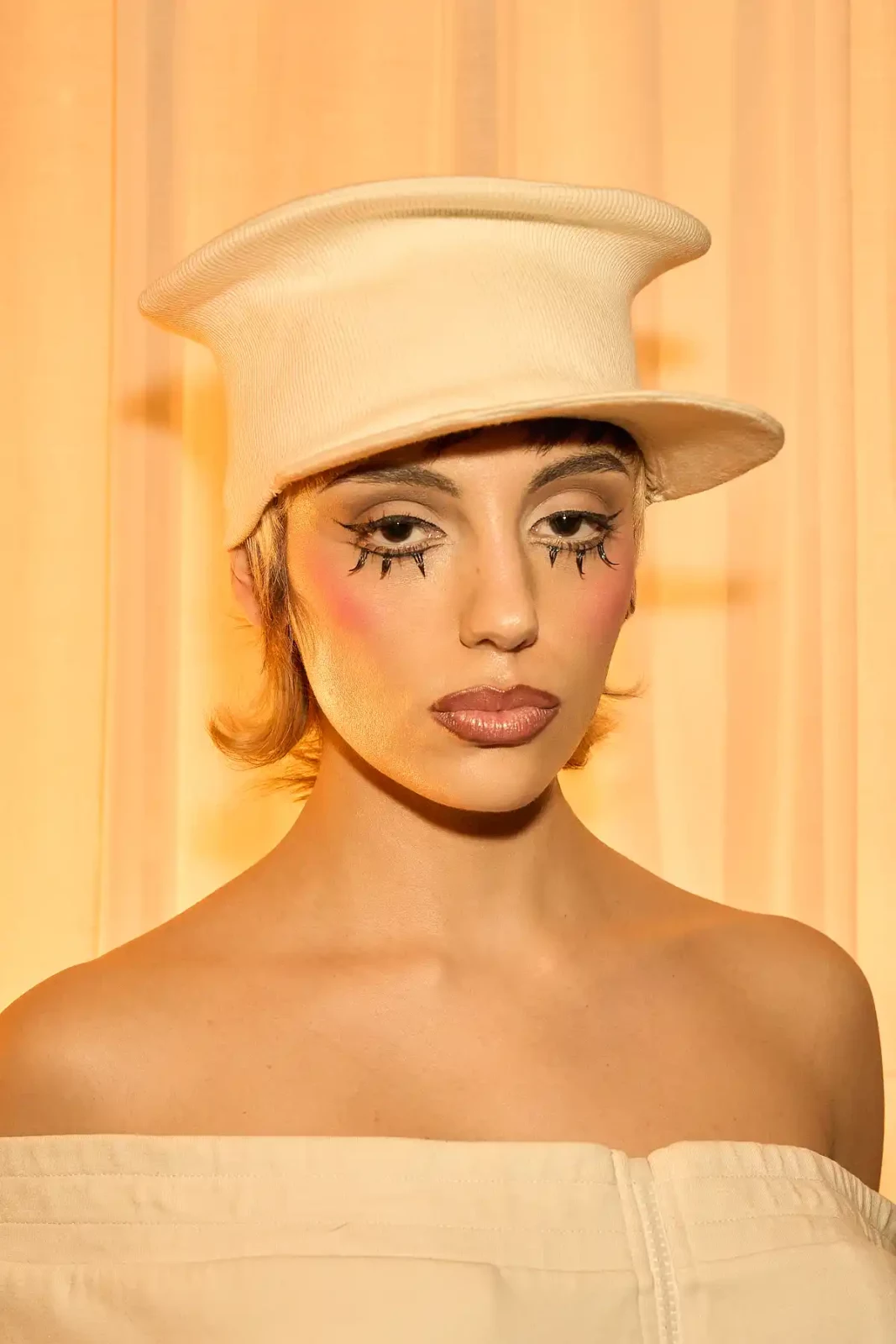 Close-up beauty portrait of a model in a white sculptural hat with dramatic eyeliner and golden lighting.