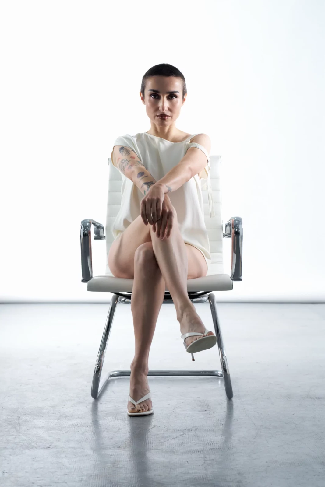 Zahara sits cross-legged on a metal chair in a white long-sleeved outfit and pointed heels.