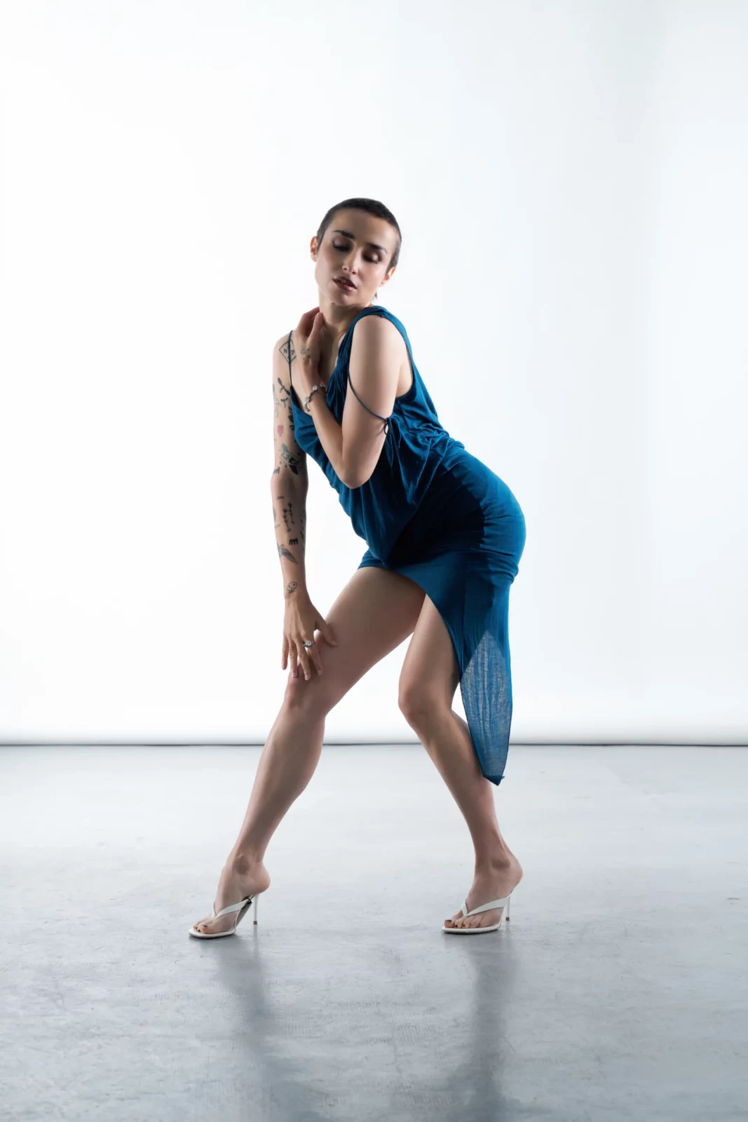 Zahara stands in a striking blue draped mini dress and high heels against a white studio backdrop.