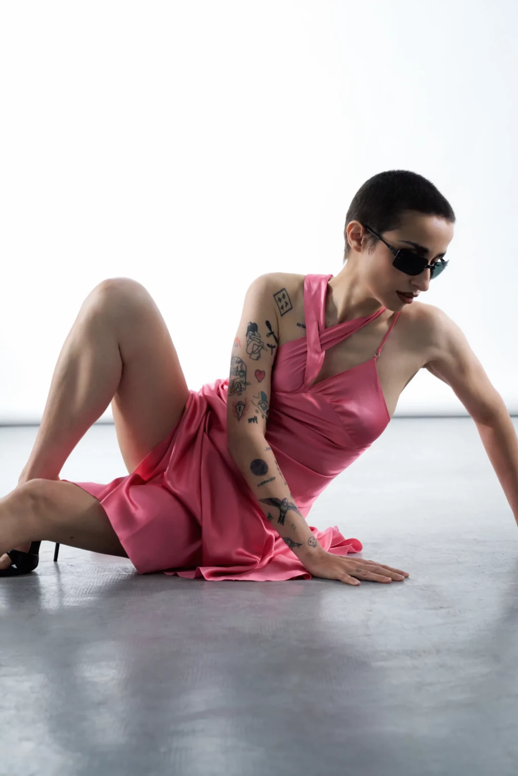 Zahara reclines on a white studio floor in a pink sculptural dress, dark sunglasses and matching heels.