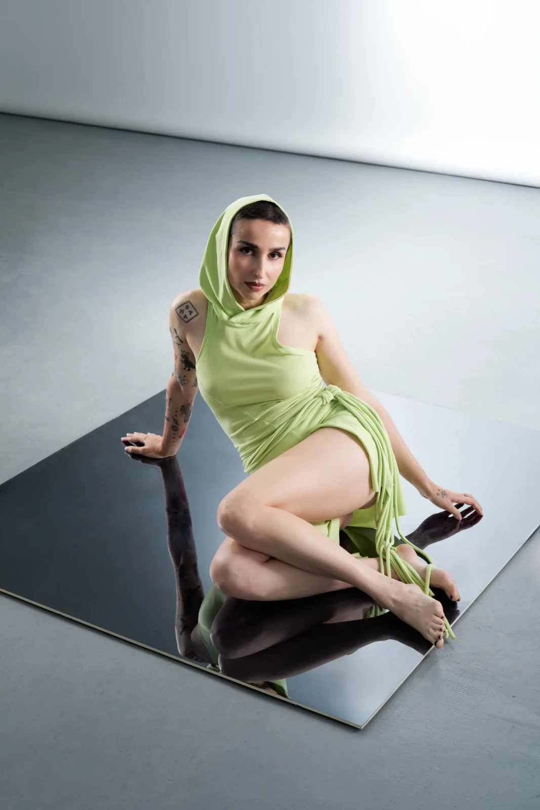 Zahara poses in a pale green hooded dress and high heels on a glossy black platform in a minimalist studio.