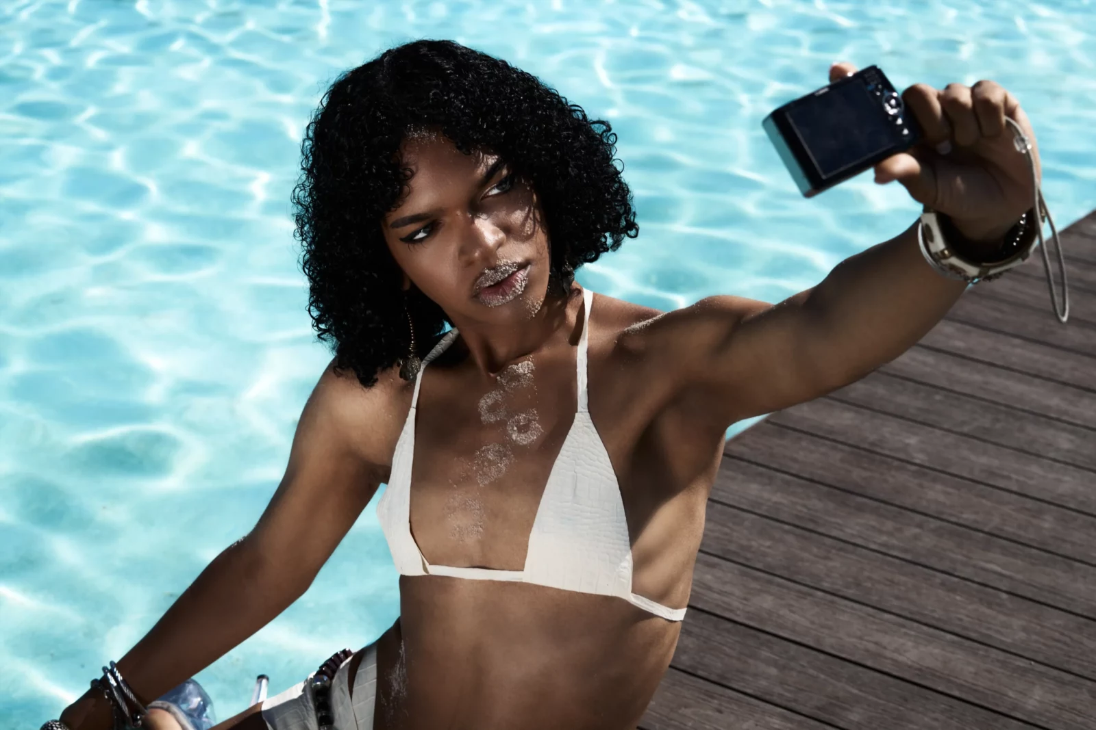 Curly-haired woman wearing a white bikini holding a small black device for a selfie beside turquoise water.