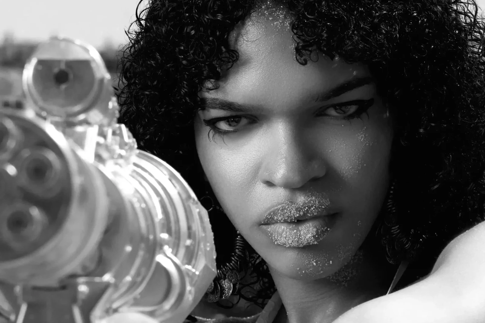 Black-and-white close-up portrait of a curly-haired woman pointing a metallic water gun at the camera.