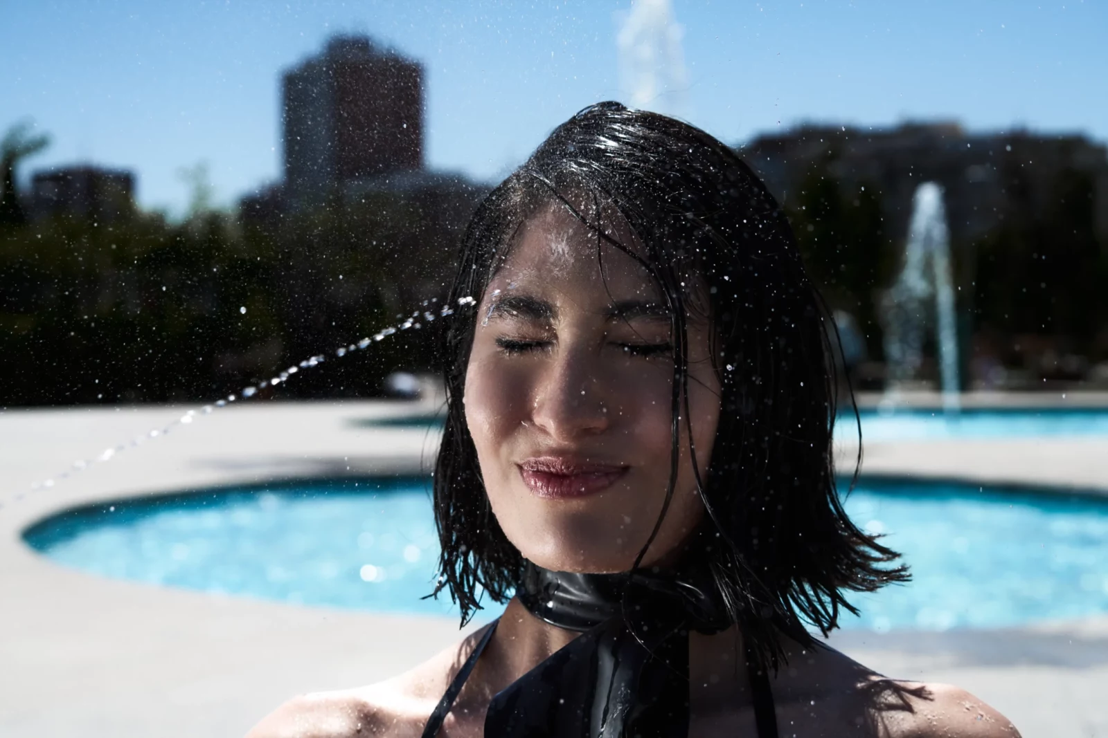 Close-up portrait of a wet-haired model in black swimwear smiling beside a blue swimming pool.