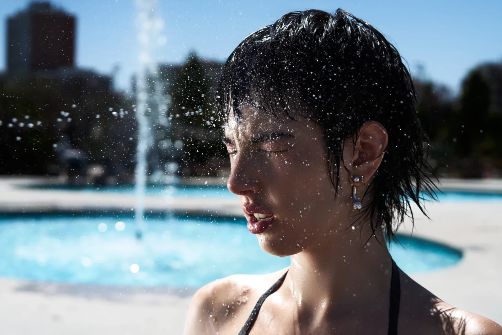 Side portrait of a short-haired model in a black bikini with water sprayed across her face.