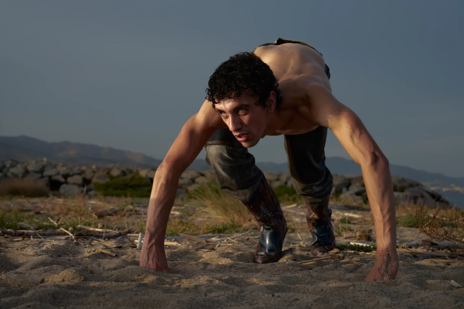 Shirtless model moves forward on all fours across an arid field with mountains in the distance.