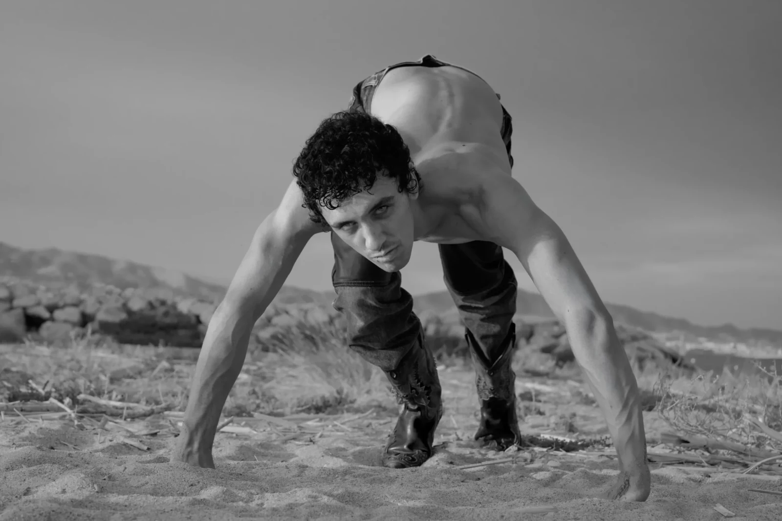 Black-and-white side view of a shirtless model bending low over rocky ground with both hands planted.
