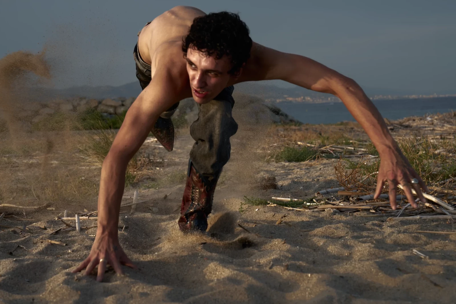 Low-angle portrait of a shirtless model crawling through dry terrain in dark, dusty trousers.