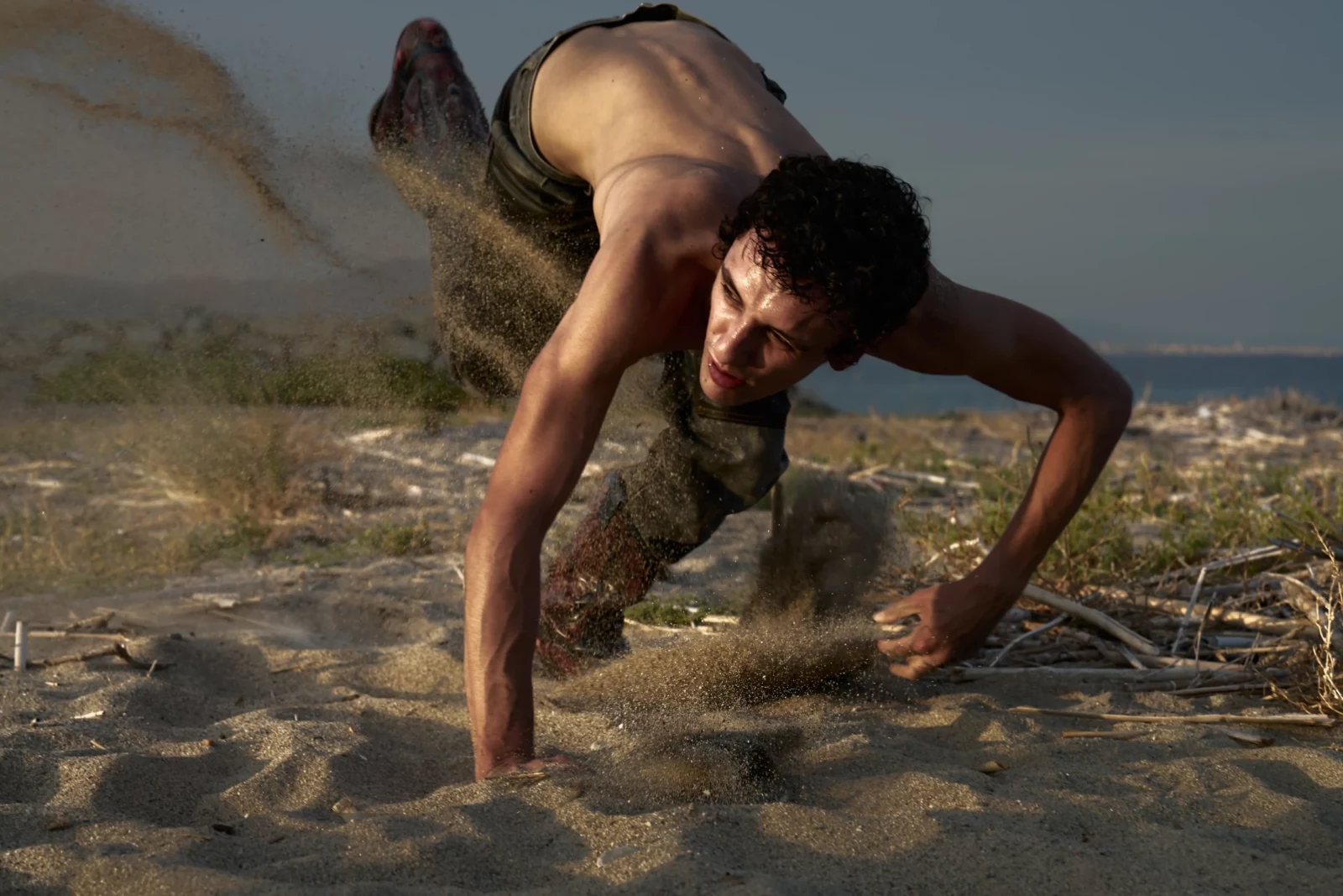 Shirtless model crawls across dusty ground in camouflage trousers under soft natural light.