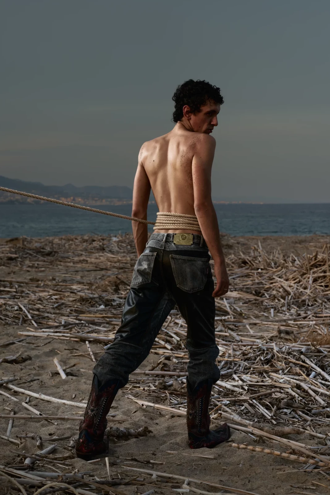 Rear view of a shirtless model walking away across a dry hillside in dark camouflage trousers.
