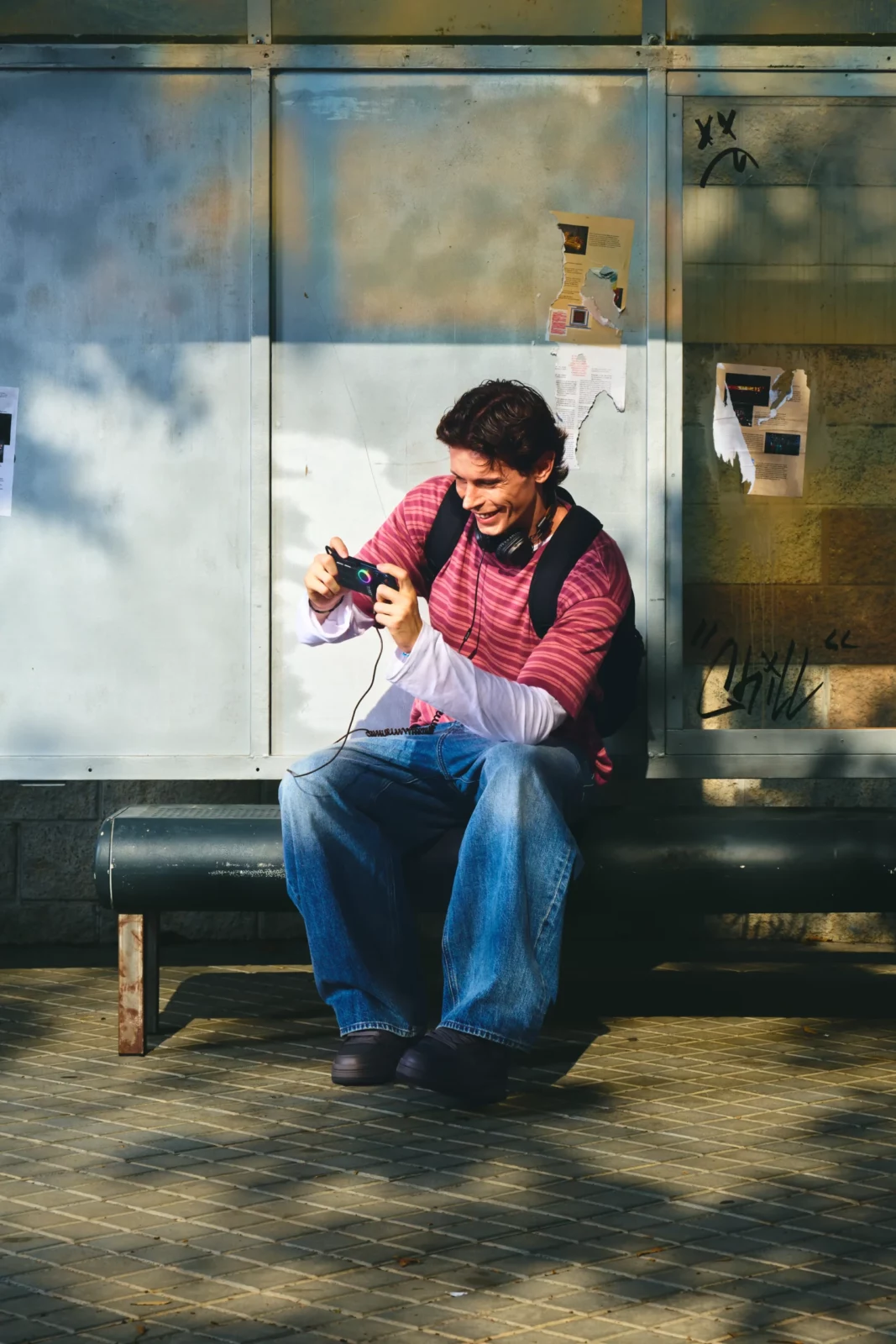 Young man in an oversized burgundy striped shirt and loose blue jeans sitting on a bench outside a glass building.
