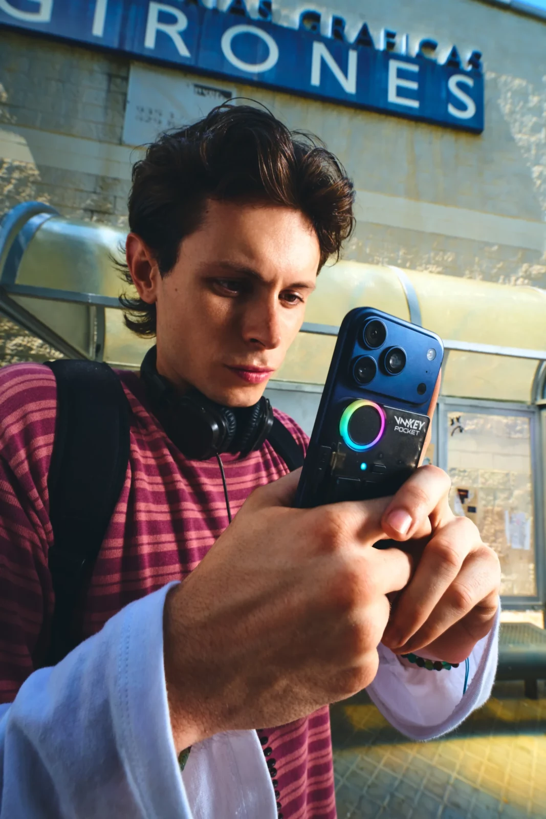 Close-up of a young man in a burgundy striped top holding a blue handheld audio device outdoors.