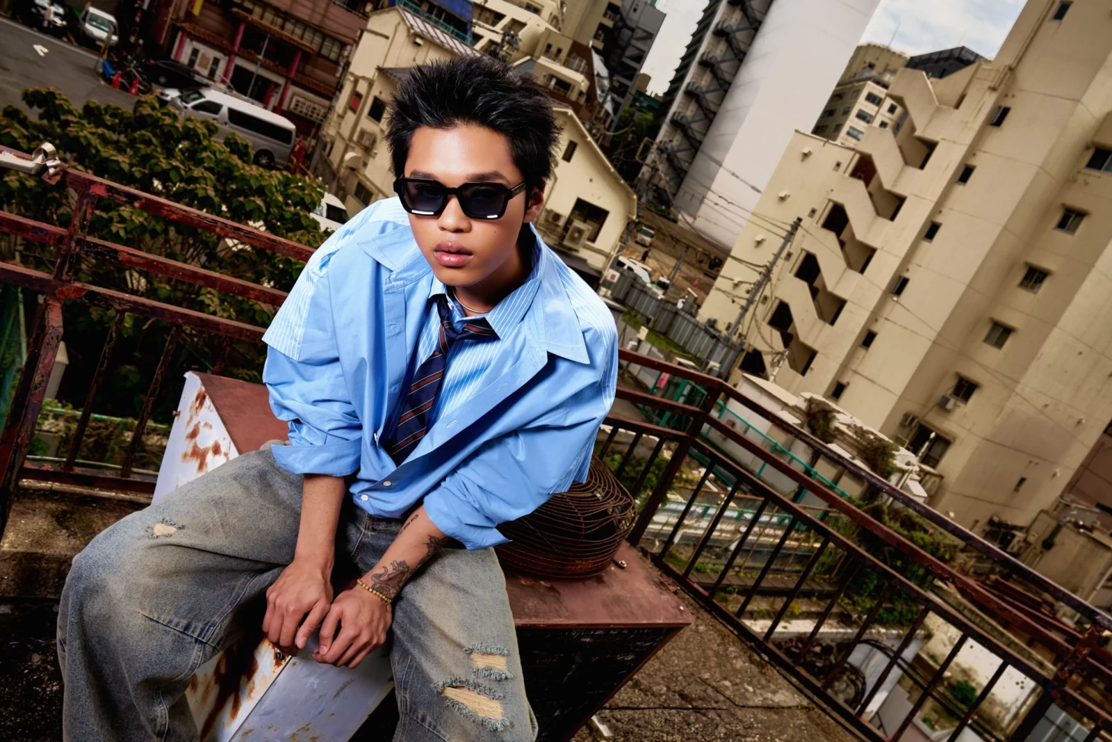 Man in sunglasses and an oversized pale blue shirt seated against a crowded Japanese city backdrop.