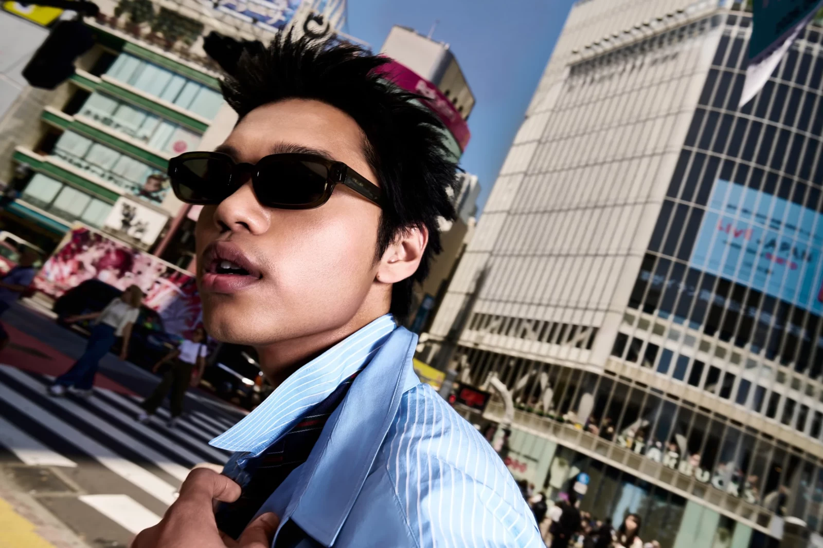 Close-up street portrait of a man in narrow black sunglasses and a blue shirt near a busy Tokyo crossing.