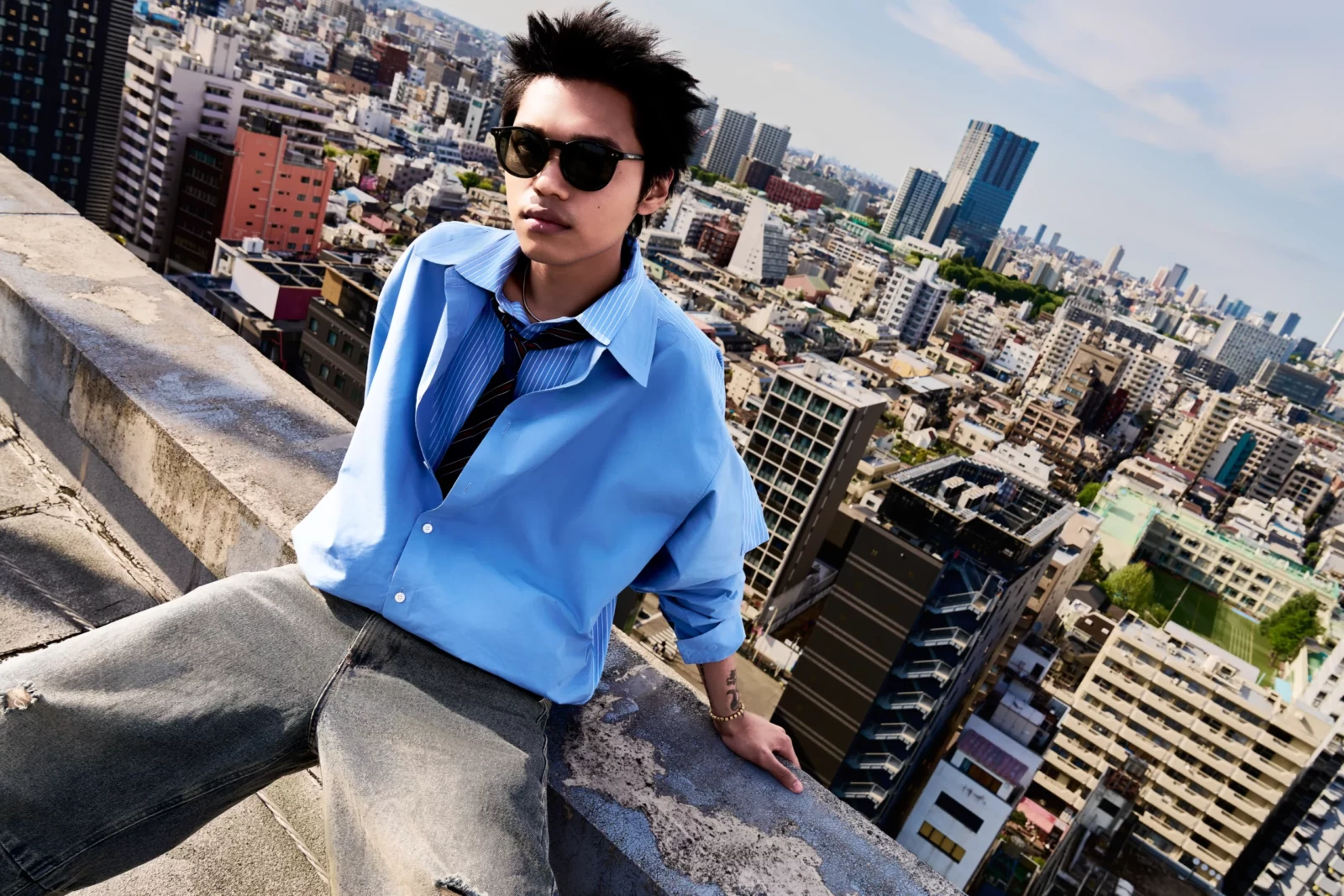 Man in dark sunglasses, a loose blue button-down shirt and gray trousers overlooking the city from a rooftop.