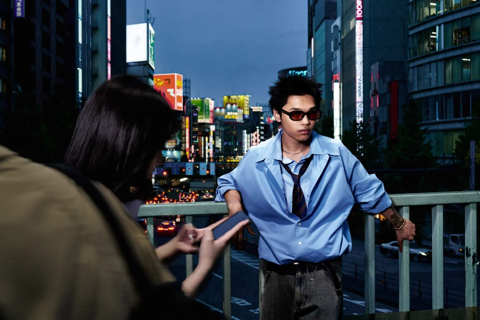 Man in black sunglasses, a loose blue shirt and tie leaning on a railing above a neon-lit Tokyo street.