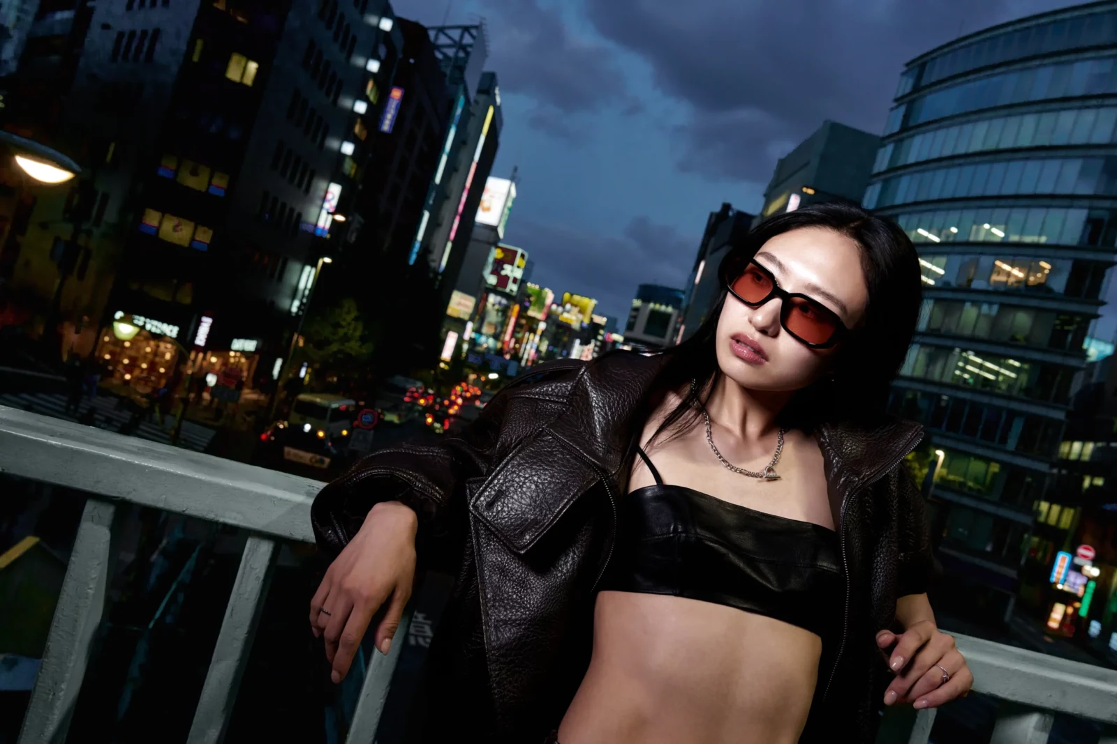 Woman in dark rectangular sunglasses, a black leather jacket and bandeau top leaning on a Tokyo bridge at dusk.