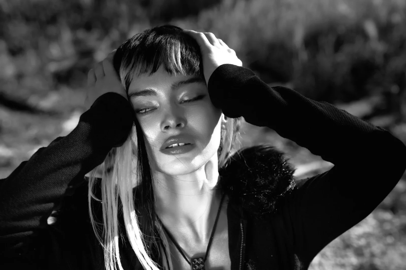 Black-and-white beauty portrait of a model with pale hair extensions holding both hands to her head.