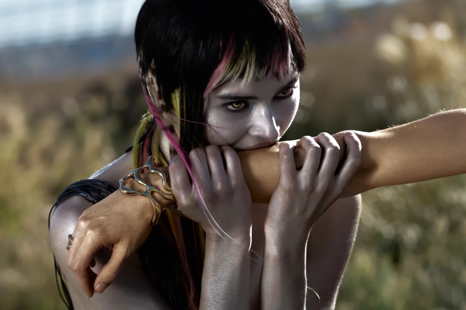 Portrait of a short-haired model with dramatic eye makeup biting their forearm against an outdoor background.