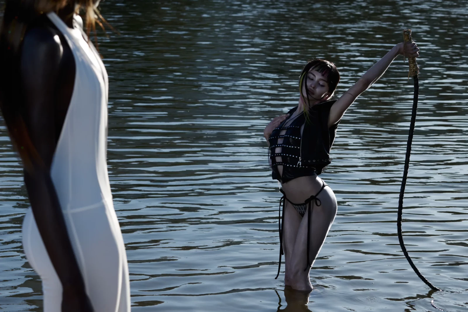 Model standing knee-deep in water wearing black swimwear and raising a long flexible object.