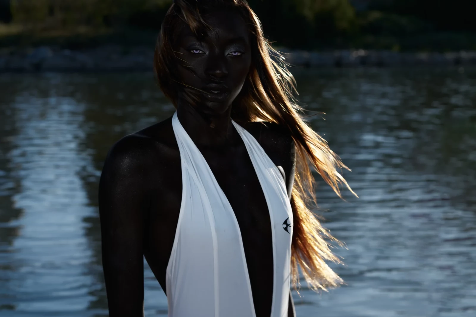 Portrait of a dark-skinned model in a sleeveless white outfit standing before shimmering water.