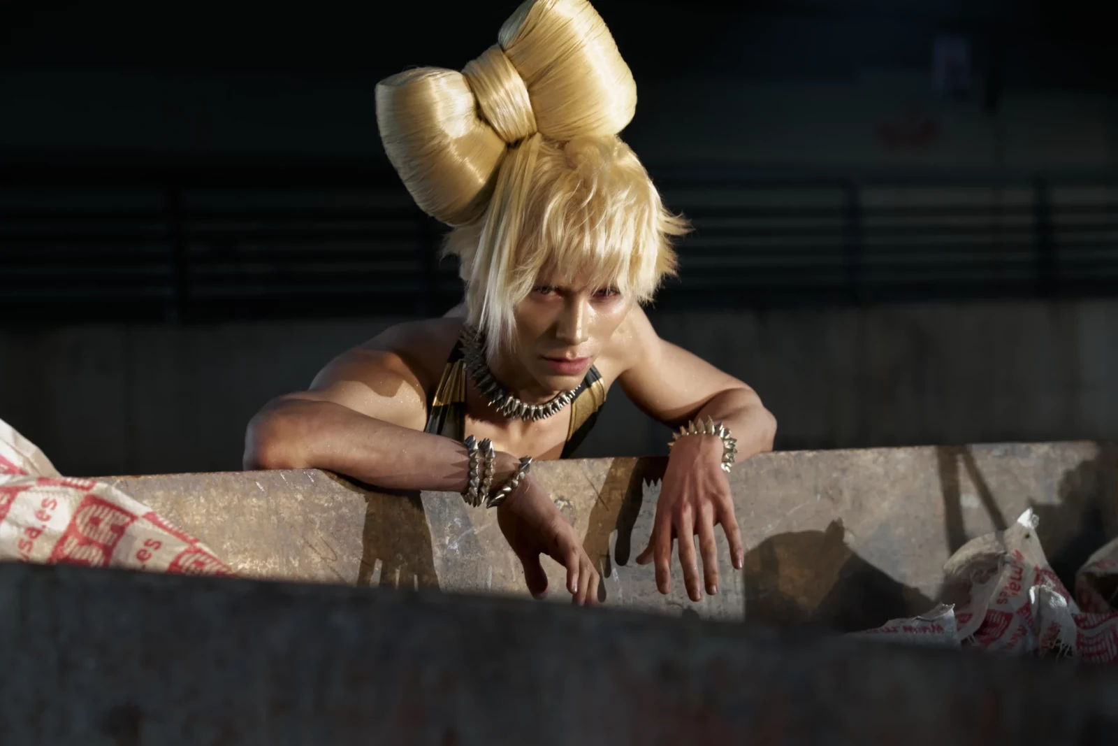 Blonde model with sculptural hair buns leaning forward over a low concrete wall at night.