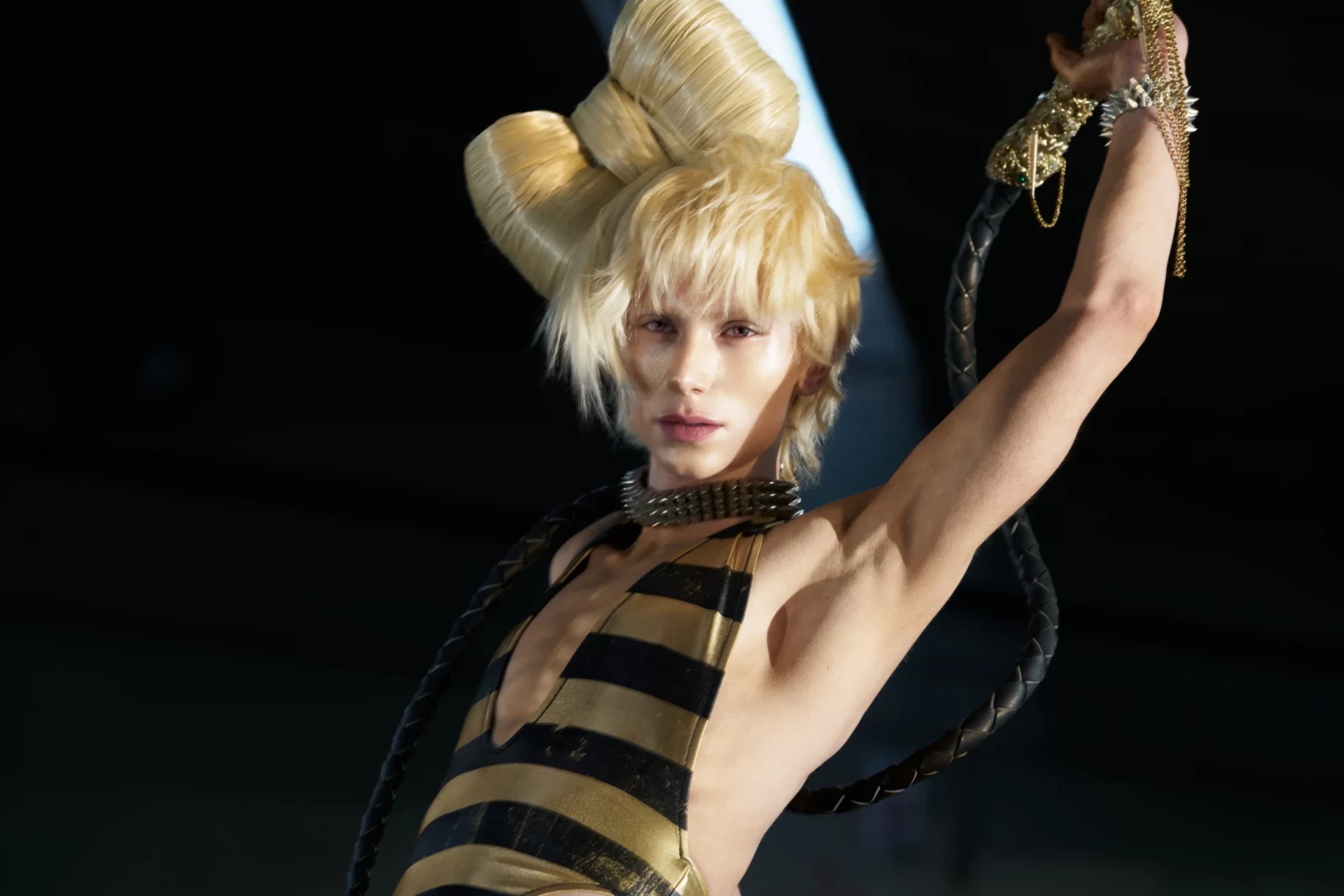 Blonde fashion model in a striped cutout outfit posing with one arm raised against a dark background.