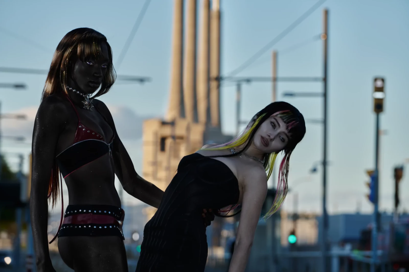 Two dark-haired fashion models in black cutout clothing posing together on a city rooftop at dusk.