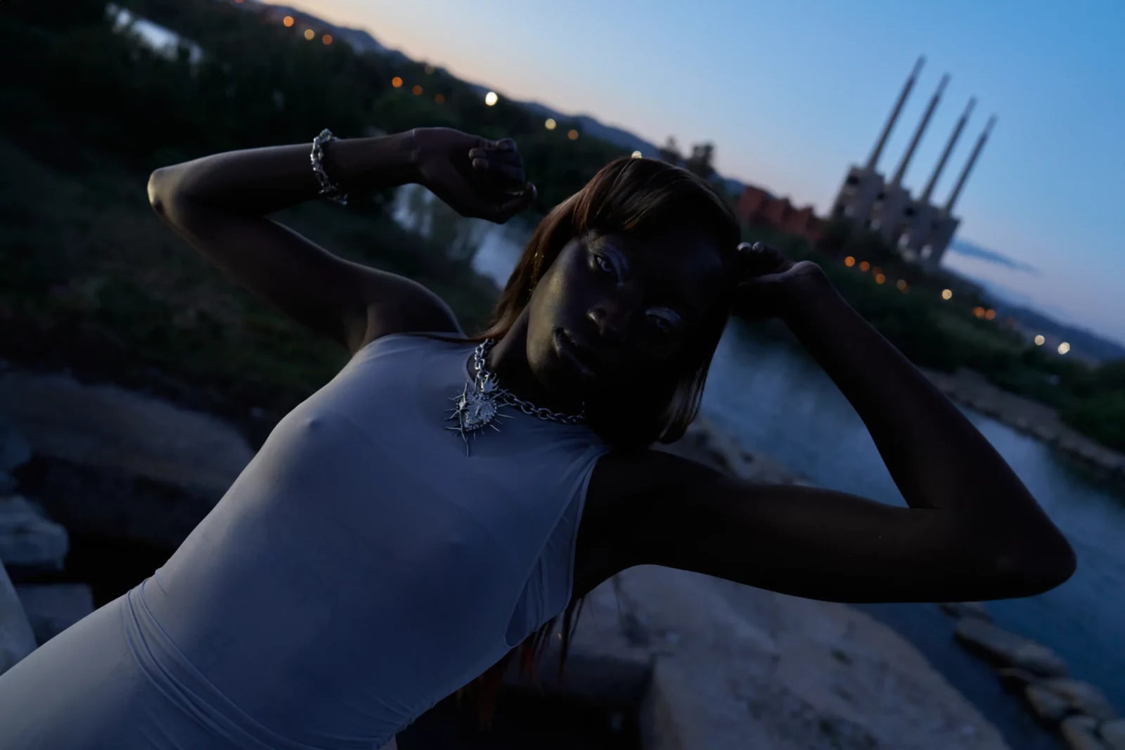 Tilted close-up of a model in a pale blue dress touching her head against a twilight waterfront.