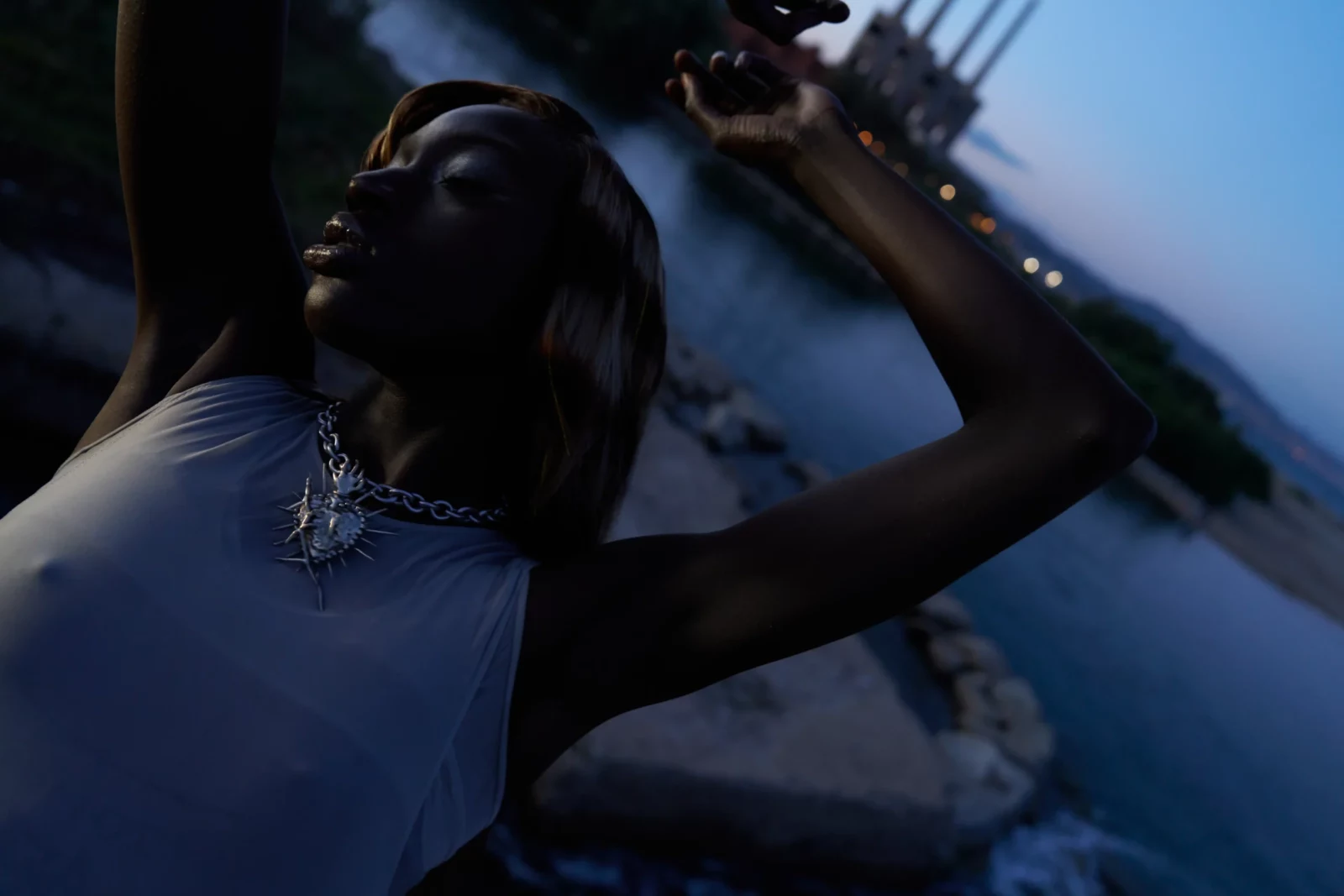 Moody close-up of a model in a pale blue sleeveless top raising one hand beside a waterfront at twilight.
